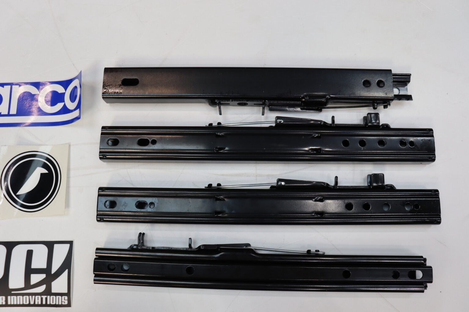 Four steel seat sliding runner rails laid out flat on a bench beside Sparco, Corbeau and PCI stickers