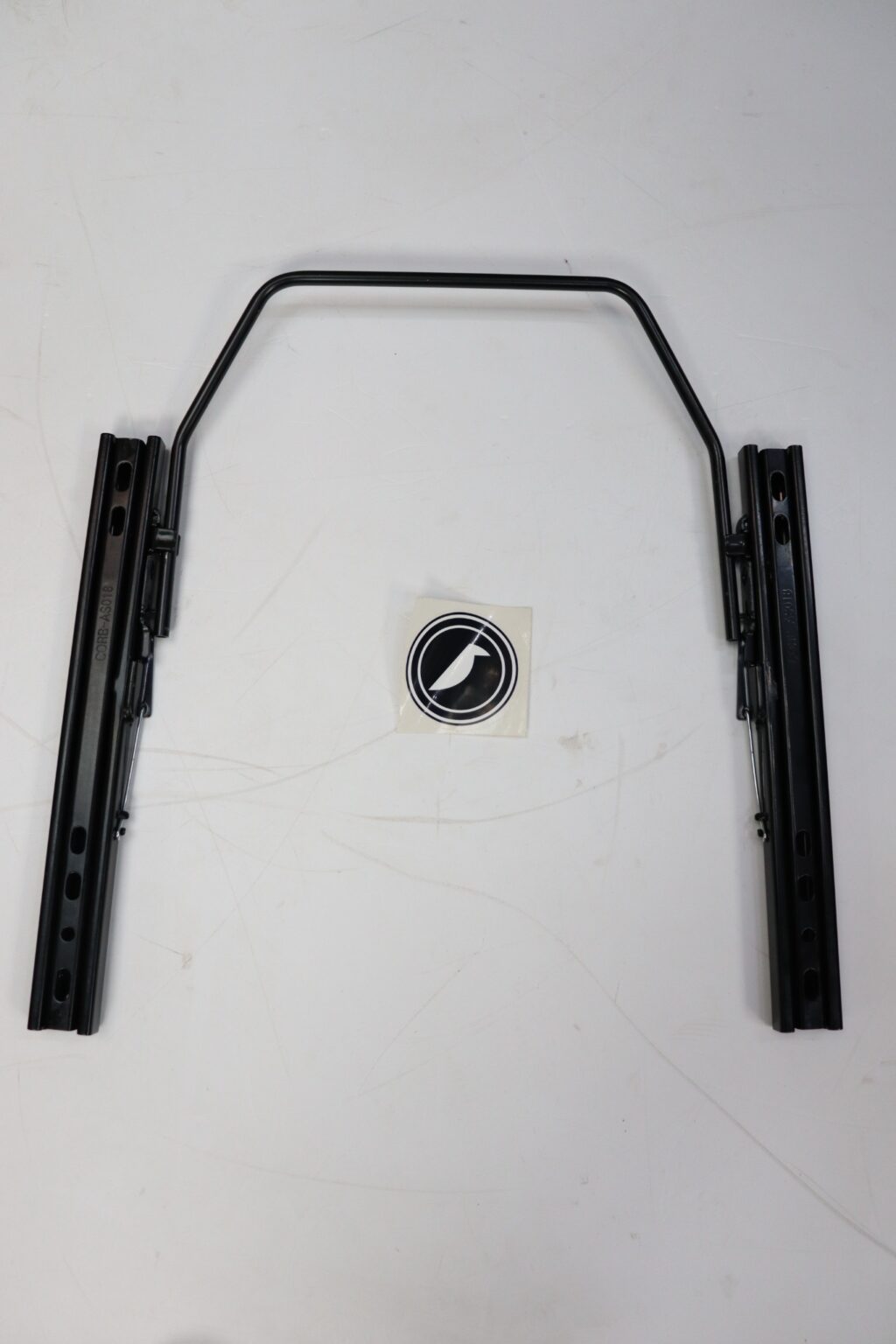 Complete Corbeau seat sliding runner kit laid out as it sits under a seat, beside the Corbeau sticker