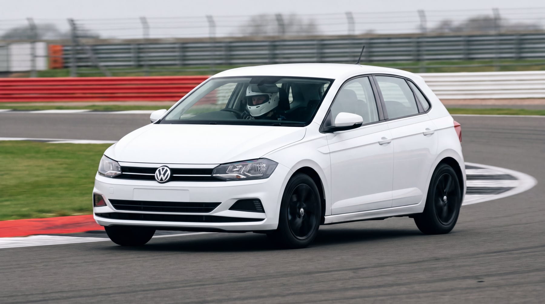 A white Volkswagen Polo Mk6 cornering on a British race circuit with a driver in a plain white crash helmet at the wheel, all four tyres gripping, no number plate fitted