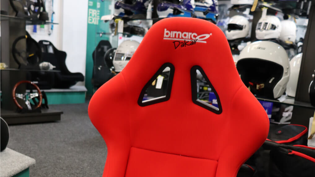 The backrest of a red velour Bimarco Dakar bucket seat in the showroom, with helmets and steering wheels on display behind