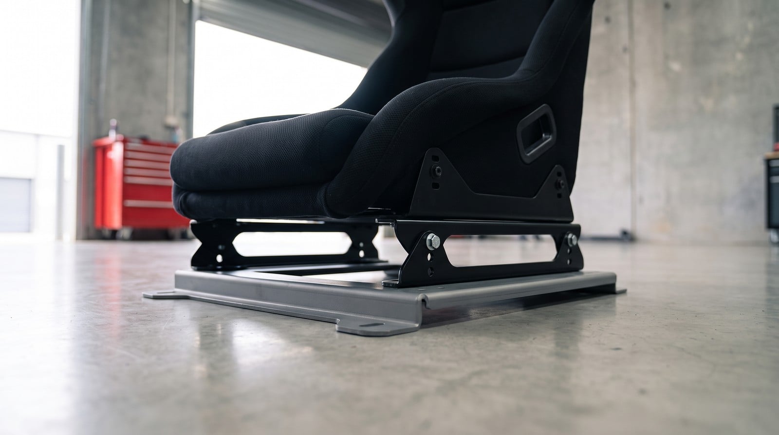 Black fixed back bucket seat on a steel sub-frame, lifted on an OMP three hole spacer rail between the sub-frame and its side mount, on a workshop floor