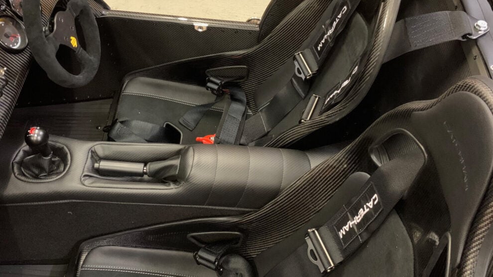 A Caterham cockpit with carbon seats and black Caterham branded harnesses, seen in a workshop