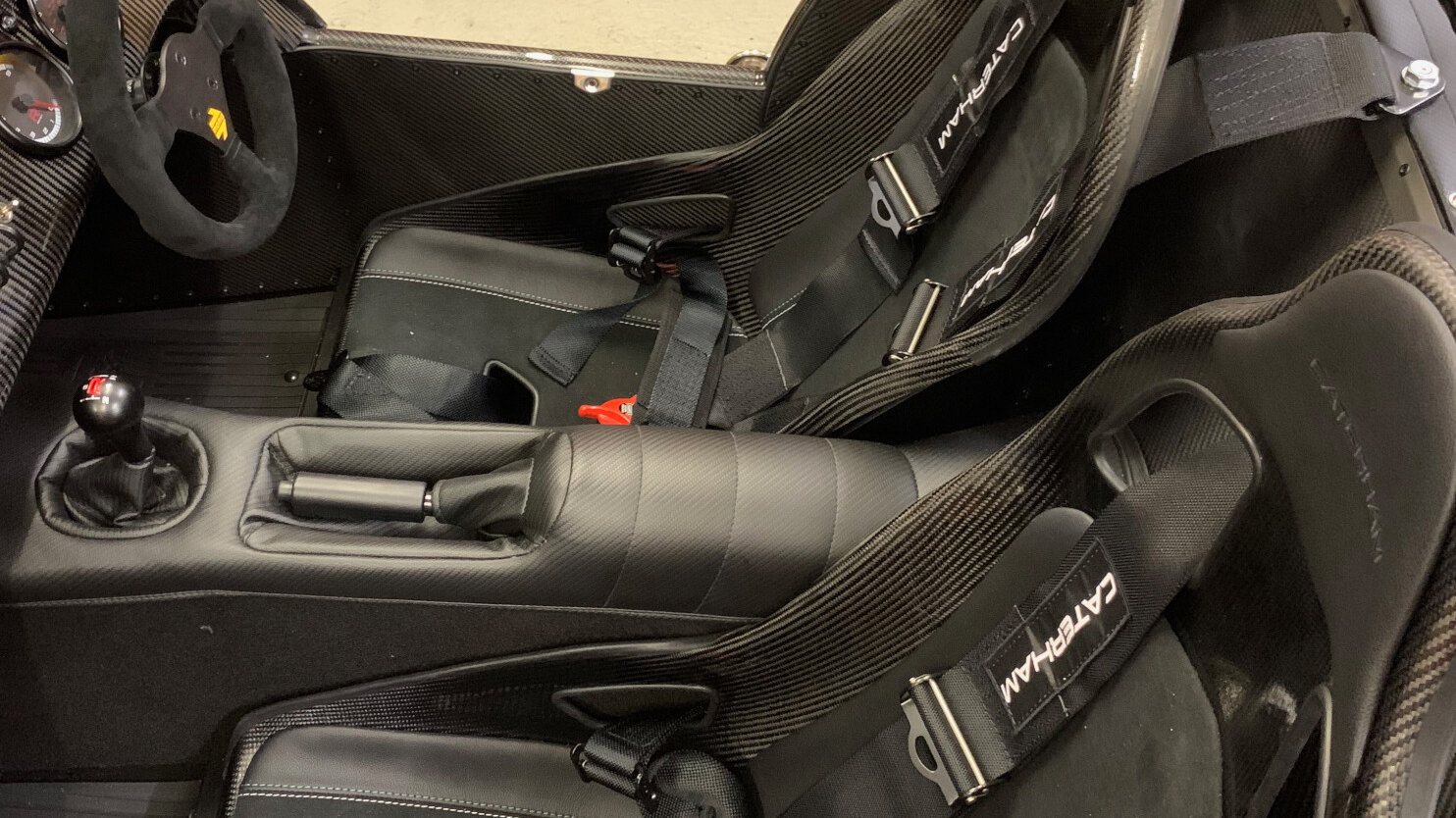 A Caterham cockpit with carbon seats and black Caterham branded harnesses, seen in a workshop