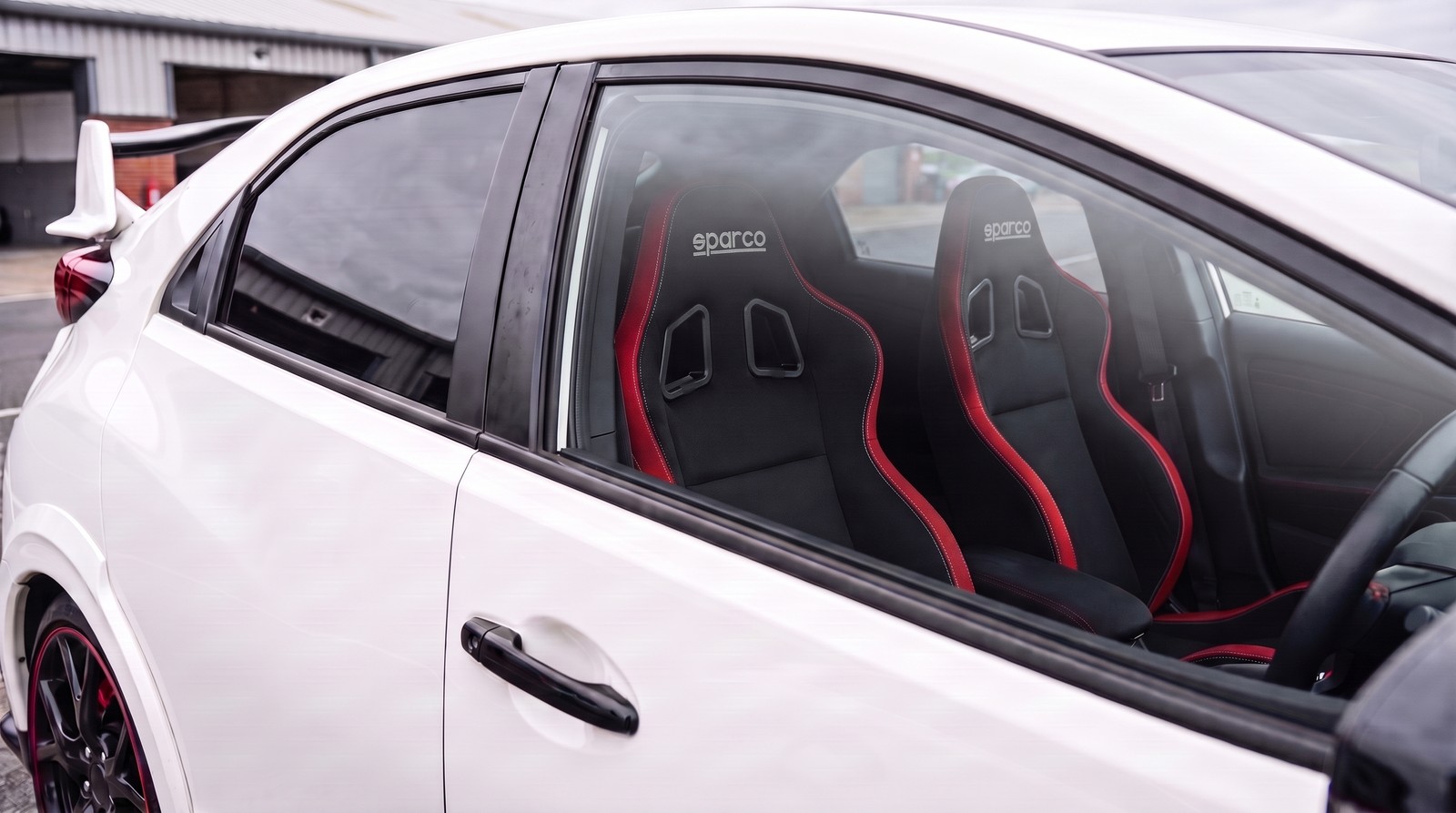 Pair of Sparco R100 reclining sports seats in black with red edge trim fitted in a Championship White 2015 Honda Civic Type R FK2, seen through the closed driver door window