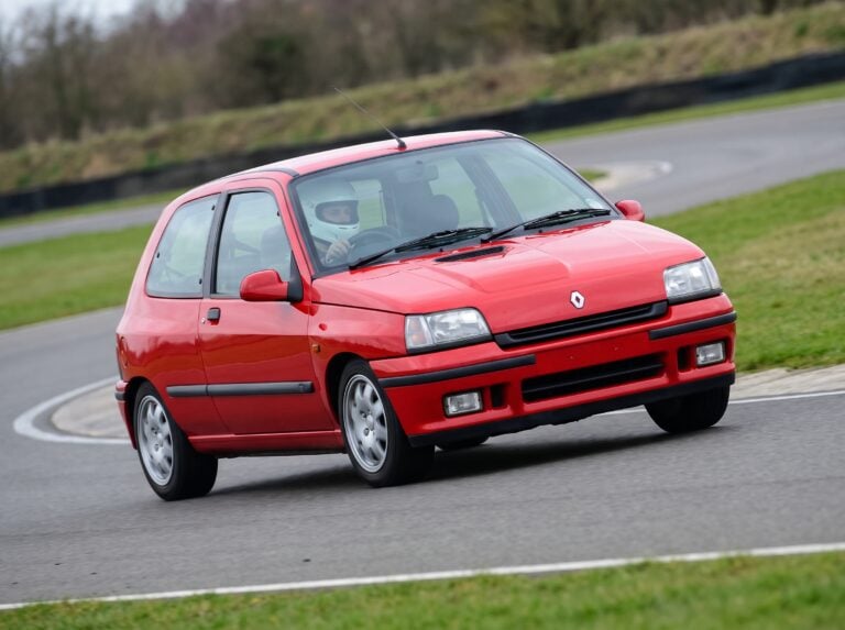 Red Renault Clio Mk1 RT hatchback cornering on a British race circuit with a helmeted driver visible through the windscreen