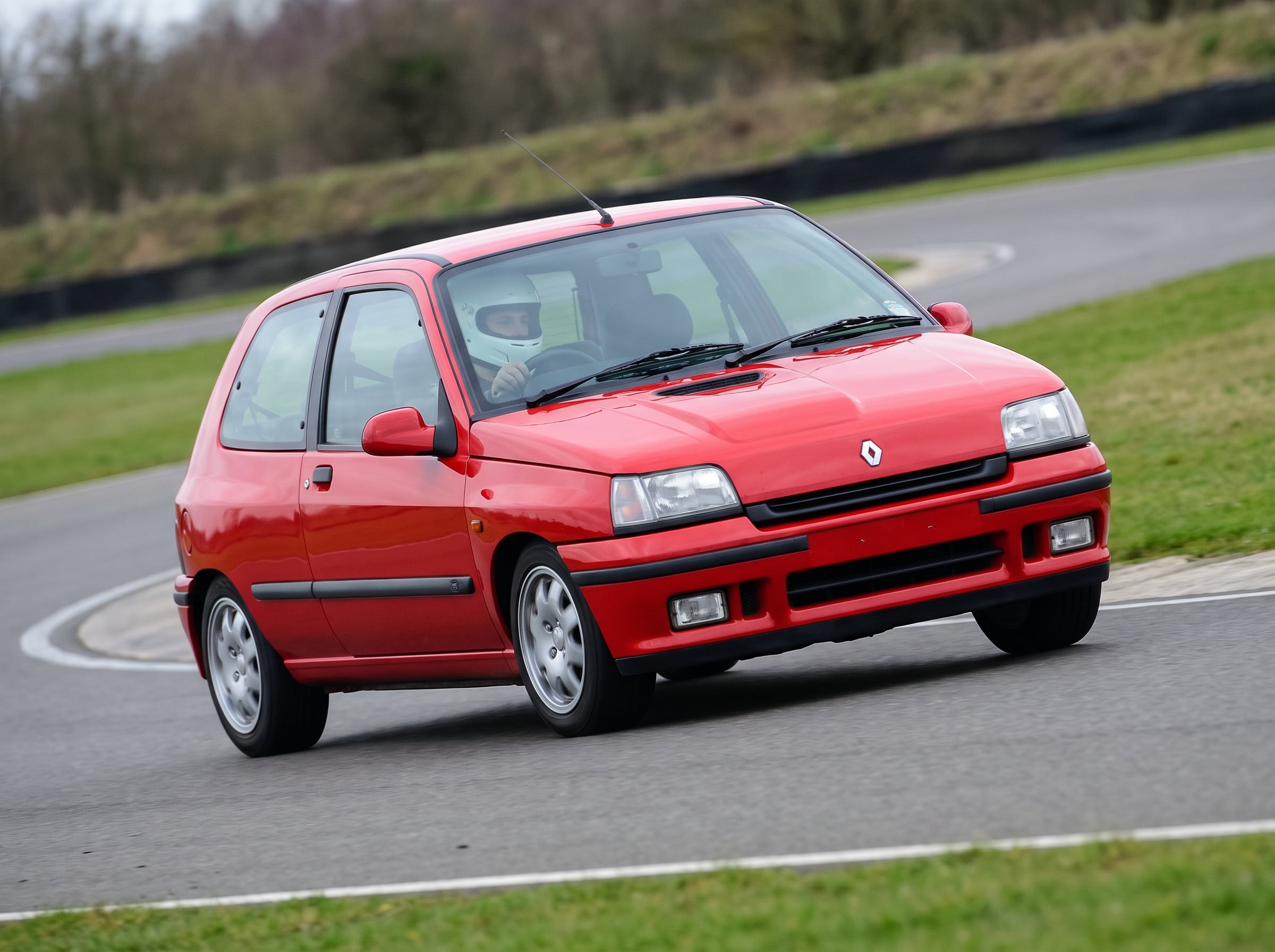 Red Renault Clio Mk1 RT hatchback cornering on a British race circuit with a helmeted driver visible through the windscreen