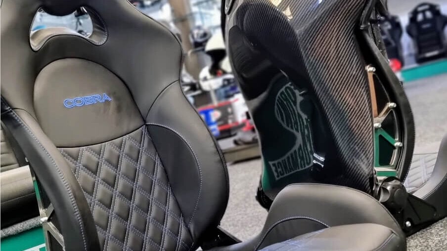 A black leather Cobra sports seat beside a carbon fibre Cobra seat shell on the showroom floor