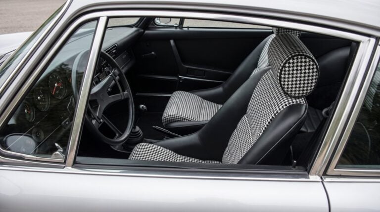Looking down into the black cabin of a silver air cooled Porsche 911 over the top of the shut driver door, showing a Cobra Classic RS seat in black with black and white houndstooth centre panels and a separate round headrest