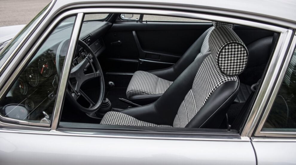 Looking down into the black cabin of a silver air cooled Porsche 911 over the top of the shut driver door, showing a Cobra Classic RS seat in black with black and white houndstooth centre panels and a separate round headrest