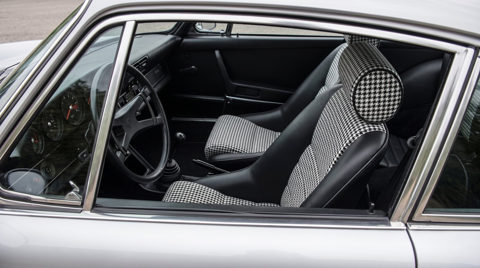 Looking down into the black cabin of a silver air cooled Porsche 911 over the top of the shut driver door, showing a Cobra Classic RS seat in black with black and white houndstooth centre panels and a separate round headrest