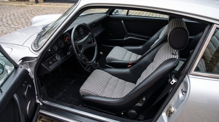 Looking down into the black cabin of a silver air cooled Porsche 911 over the top of the shut driver door, showing a Cobra Classic RS seat in black with black and white houndstooth centre panels and a separate round headrest