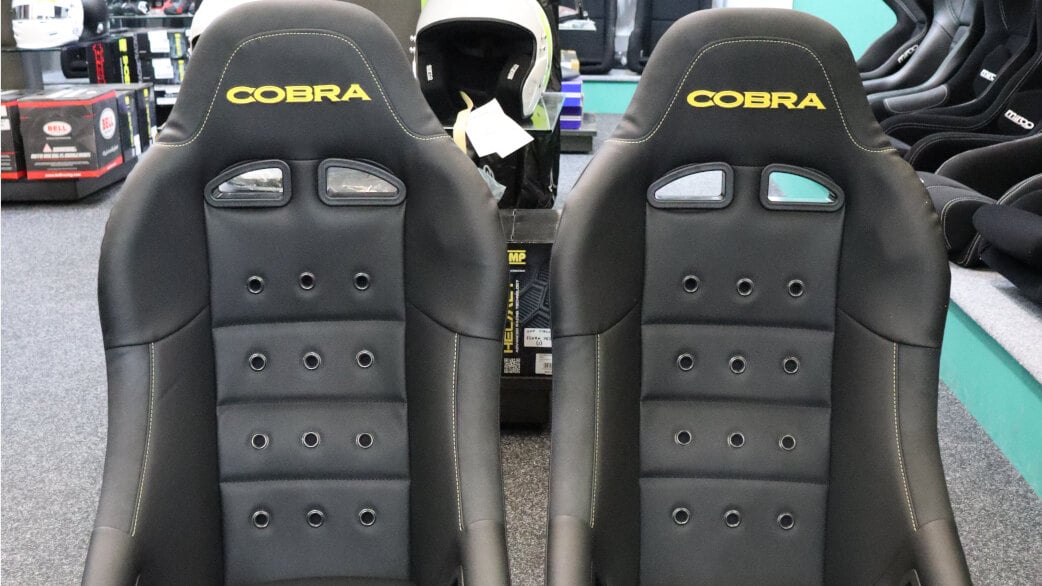 A pair of black vinyl Cobra Clubman 40th Edition bucket seats with yellow Cobra logos, yellow stitching and anthracite eyelets on the showroom floor