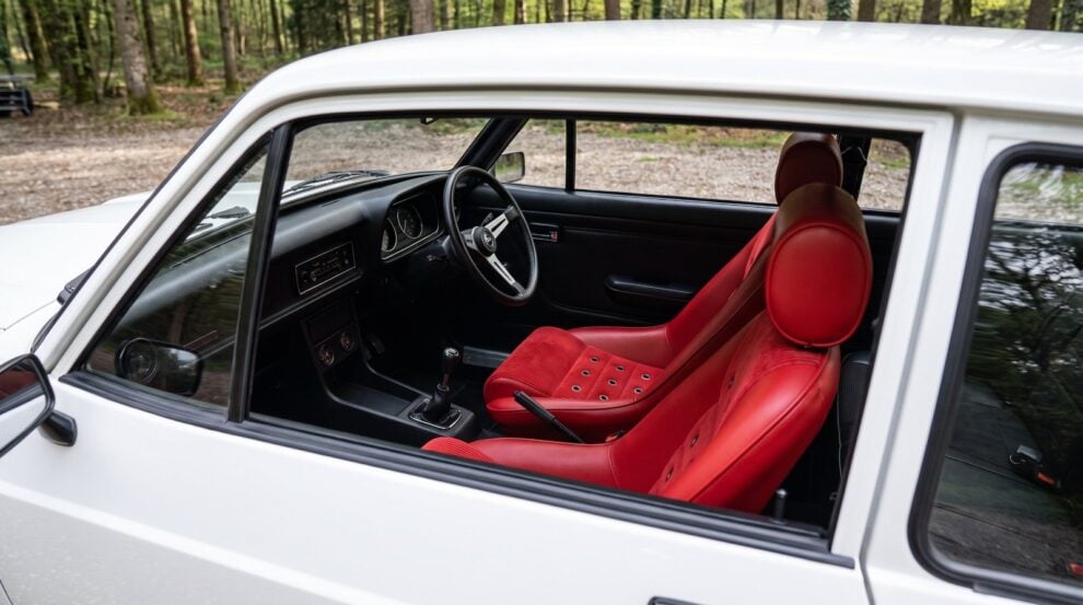 Looking down into the black cabin of a white Ford Escort Mk2 over the top of the shut driver door, showing a Cobra GT3 Historic fixed back seat in red vinyl with red corduroy centre panels and a grid of punched eyelets in the backrest
