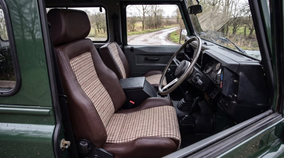Looking down into the cabin of a dark green Land Rover Defender 90 over the top of the shut driver door, showing a Cobra Le Mans reclining seat in dark brown vinyl with oatmeal and brown Harris Tweed houndstooth centre panels