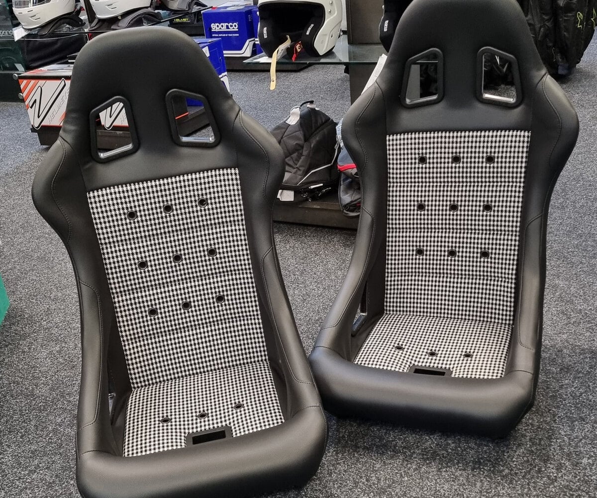 A pair of black vinyl Cobra Monaco 40th bucket seats with black and white houndstooth centre panels and eyelets, on the showroom floor in front of helmet displays