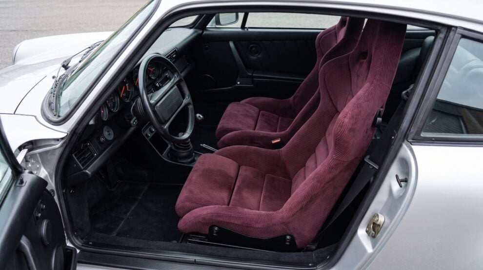 Looking down into the black cabin of a silver air cooled Porsche 911 over the top of the shut driver door, showing a Cobra Nogaro Circuit bucket seat trimmed all over in burgundy corduroy with harness slots cut through the shoulders