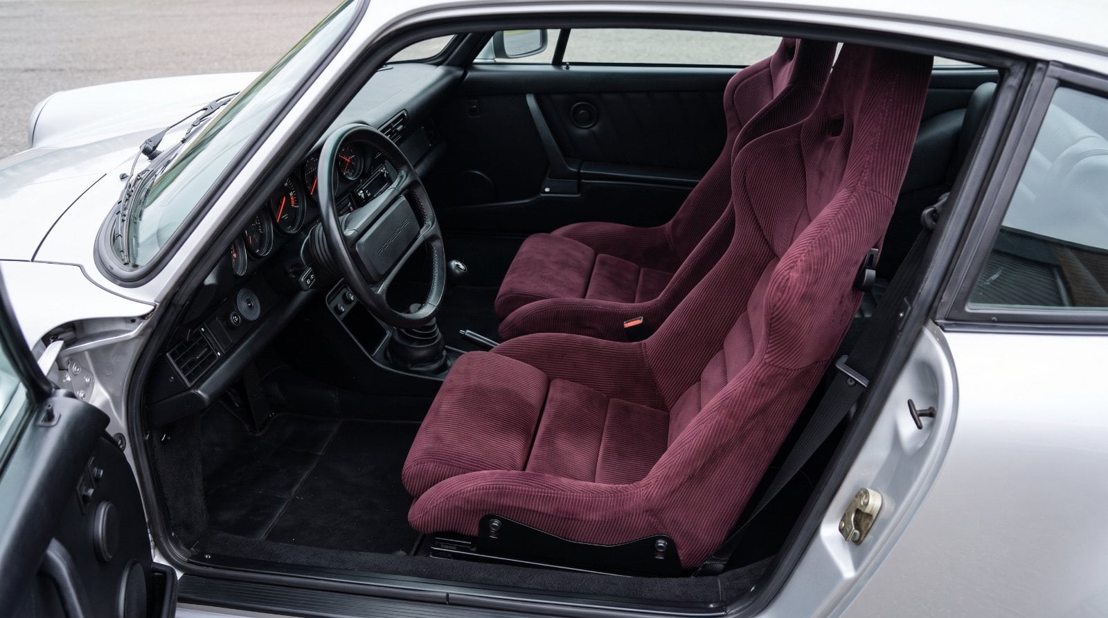 Looking down into the black cabin of a silver air cooled Porsche 911 over the top of the shut driver door, showing a Cobra Nogaro Circuit bucket seat trimmed all over in burgundy corduroy with harness slots cut through the shoulders