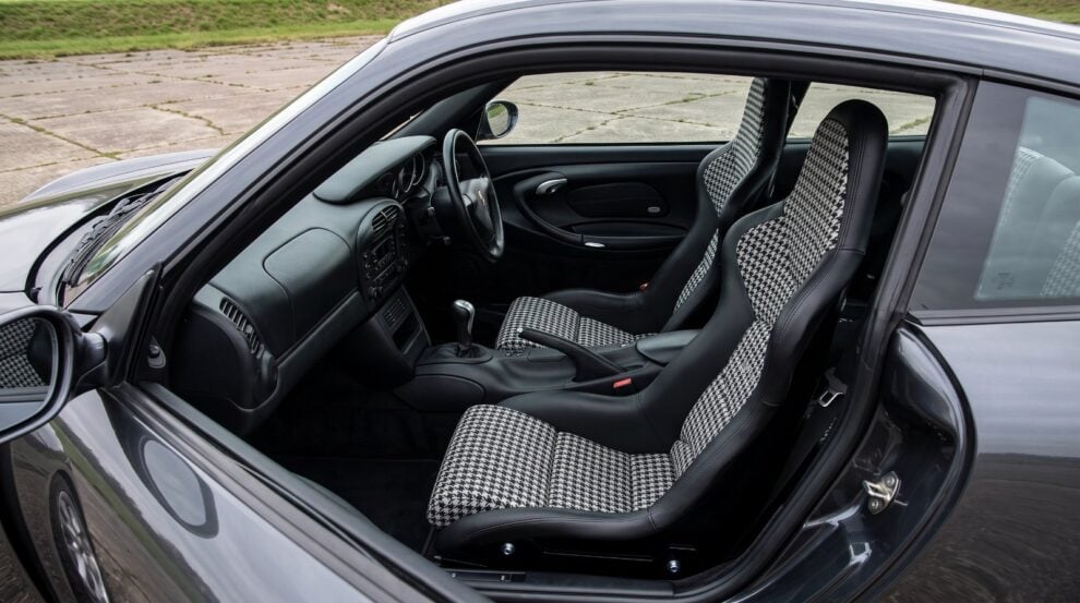 Looking down into the black leather cabin of a dark grey modern Porsche 911 over the top of the shut driver door, showing a Cobra Nogaro Street one piece fixed back bucket seat with black and white houndstooth centre panels
