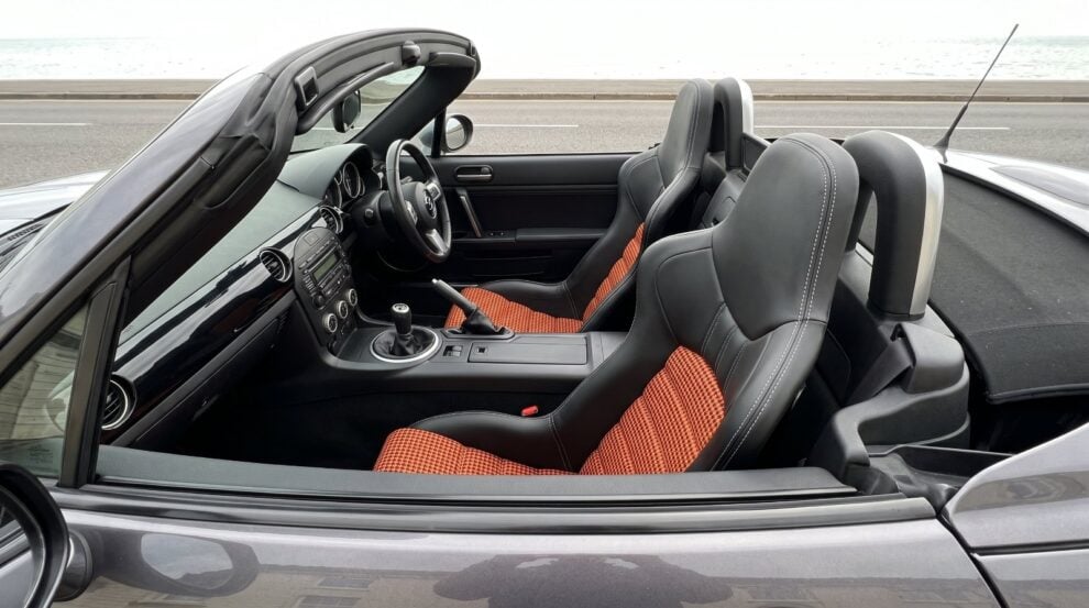 Looking down into the open cockpit of a dark grey Mazda MX-5 Mk3 over the top of the shut driver door, showing a Cobra Nogaro Street bucket seat in black vinyl with orange and black pepita houndstooth centre panels and white stitching