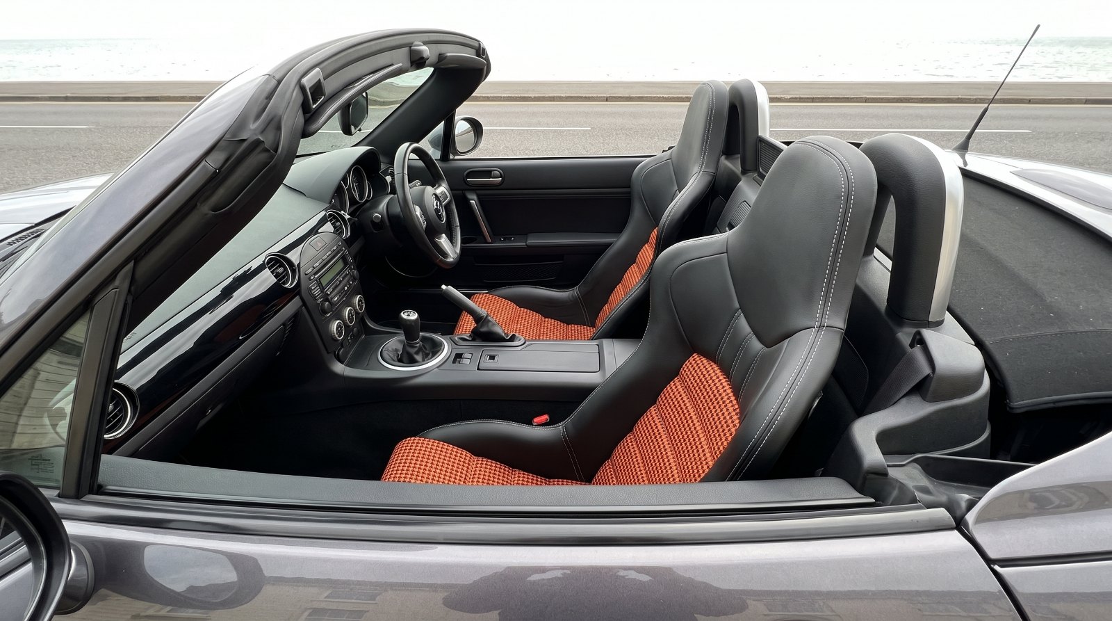 Looking down into the open cockpit of a dark grey Mazda MX-5 Mk3 over the top of the shut driver door, showing a Cobra Nogaro Street bucket seat in black vinyl with orange and black pepita houndstooth centre panels and white stitching
