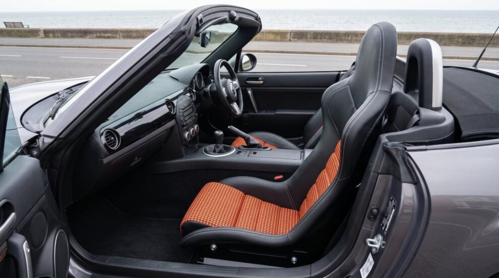 Close view of a Cobra Nogaro Street fixed back bucket seat fitted in a Mazda MX-5 Mk3, in black vinyl with orange and black pepita houndstooth centre panels and white stitching