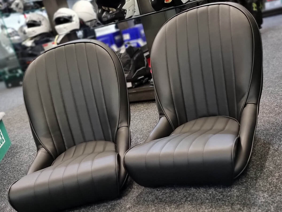 A pair of black Cobra Roadster SS historic bucket seats with vertical pleats on the showroom carpet, with helmets on display behind