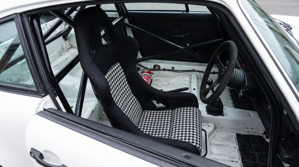 Looking down into the stripped cabin of a caged competition car over the top of the shut driver door, showing a Cobra Suzuka Pro-Fit FIA bucket seat with harness slots cut through the shoulders and black and white houndstooth centre panels