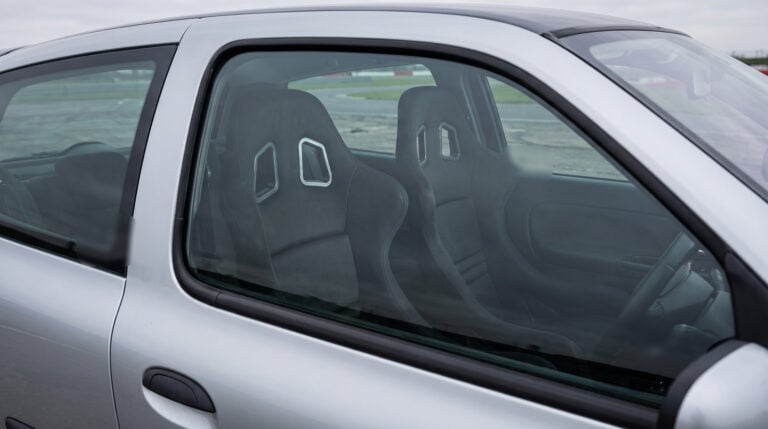A pair of black Cobra Suzuka T bucket seats seen through the closed driver door window of a silver 2003 Renault Clio Mk2 in a race circuit paddock