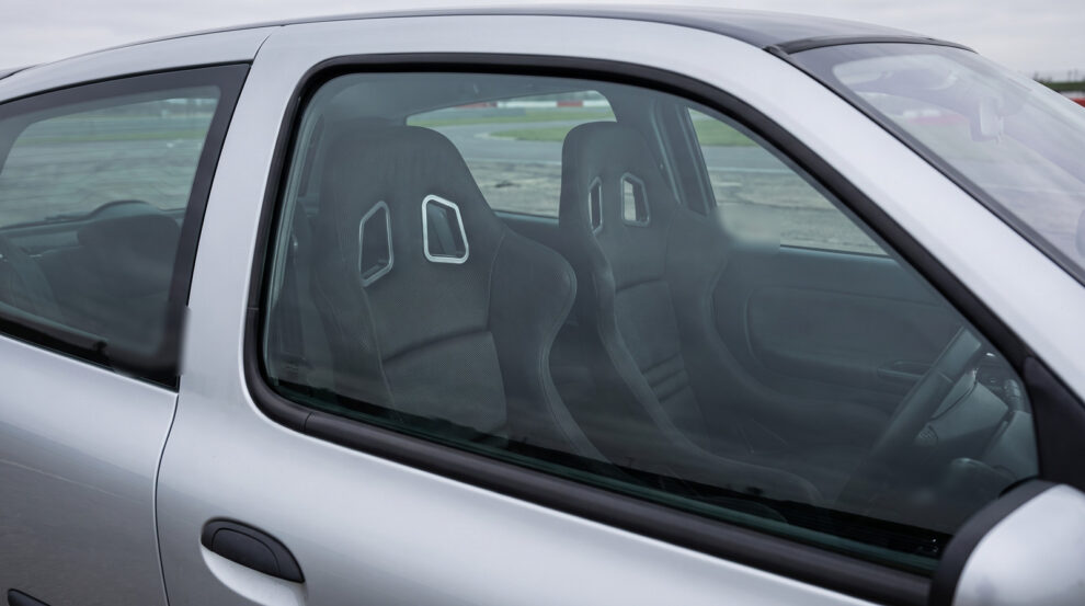 A pair of black Cobra Suzuka T bucket seats seen through the closed driver door window of a silver 2003 Renault Clio Mk2 in a race circuit paddock