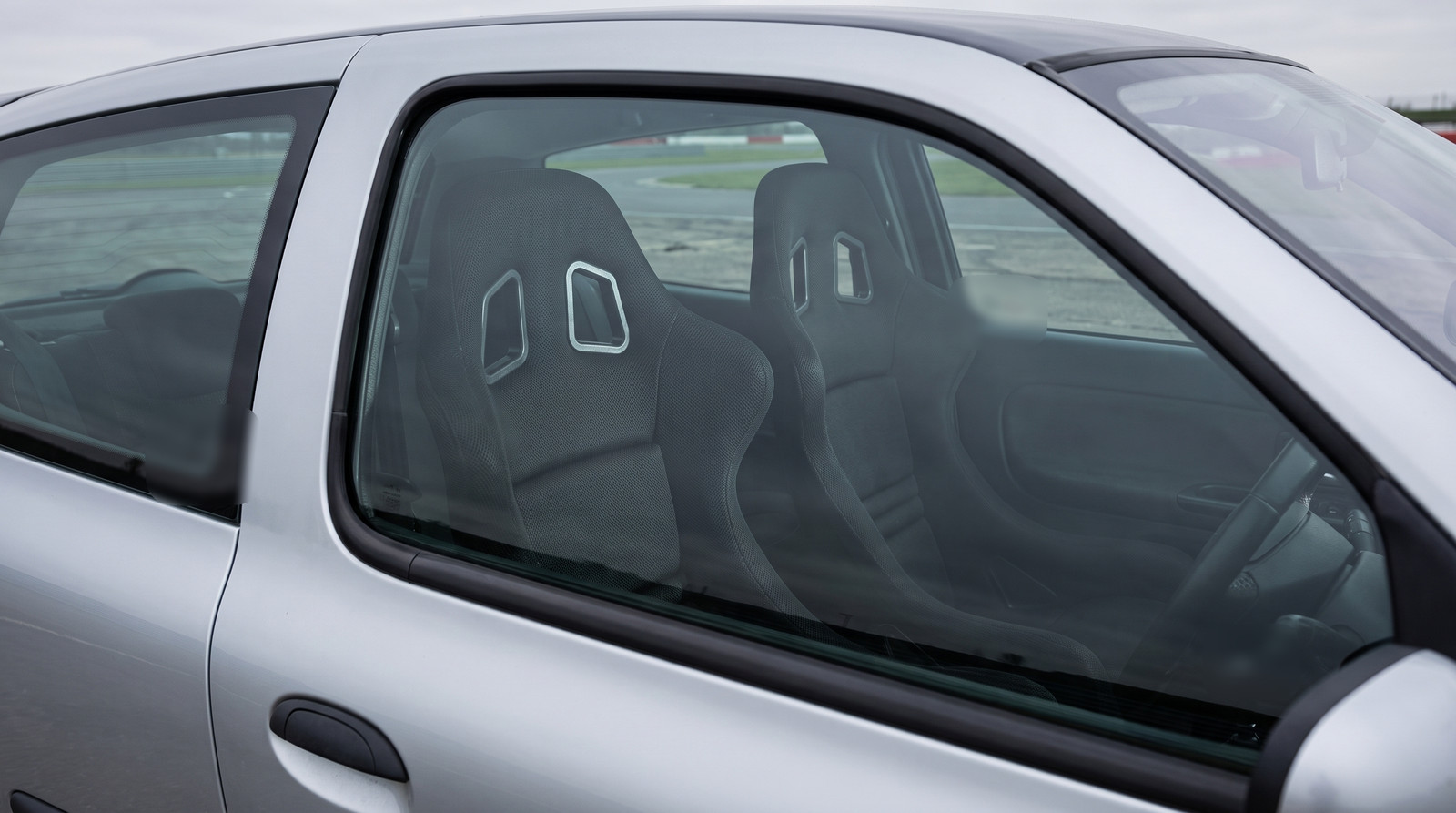 A pair of black Cobra Suzuka T bucket seats seen through the closed driver door window of a silver 2003 Renault Clio Mk2 in a race circuit paddock