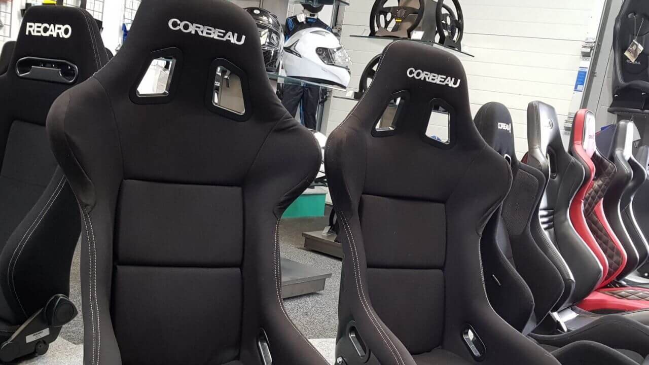 Two black Corbeau Clubsport bucket seats on the GSM Performance showroom plinth, with helmets and steering wheels on the shelves behind
