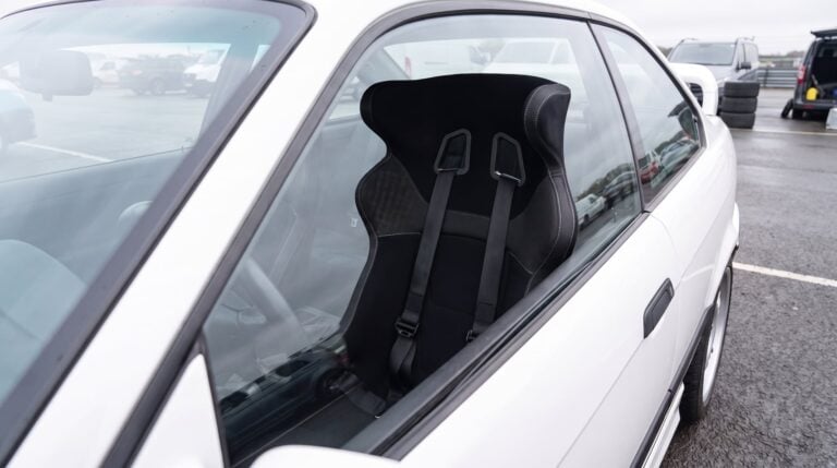 A black Corbeau Revenge fixed back bucket seat with a harness fitted in a white BMW E36 track day car, seen through the closed driver door window in a race circuit paddock