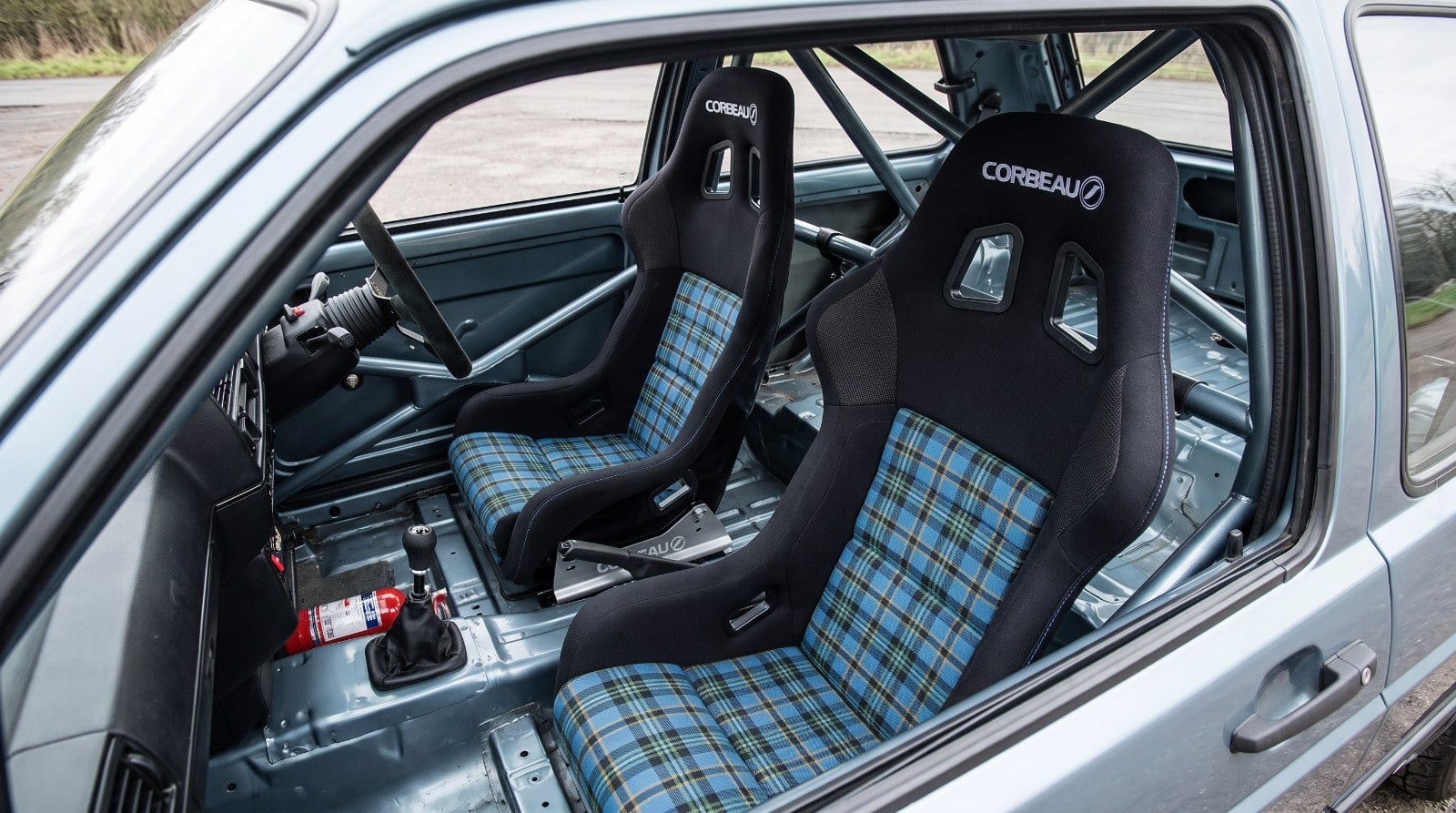 Two Corbeau Tartan Edition FIA bucket seats with black shells and blue tartan centre panels, mounted on alloy side mounts in a caged competition car, seen through the wound down driver window over the top of the shut door