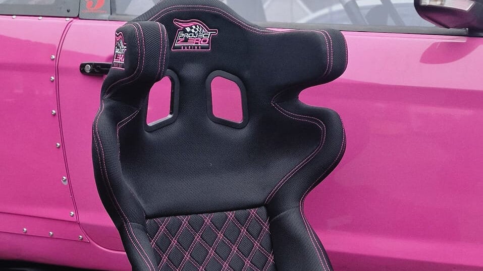 A customised black Mirco RS7 bucket seat with pink stitching and embroidered logos in front of a pink race car