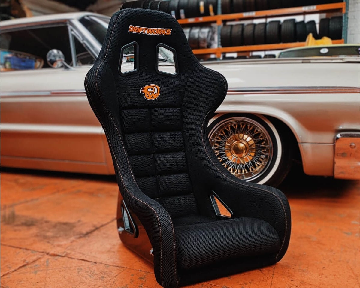 A black Driftworks bucket seat with orange stitching standing on an orange workshop floor in front of a classic car