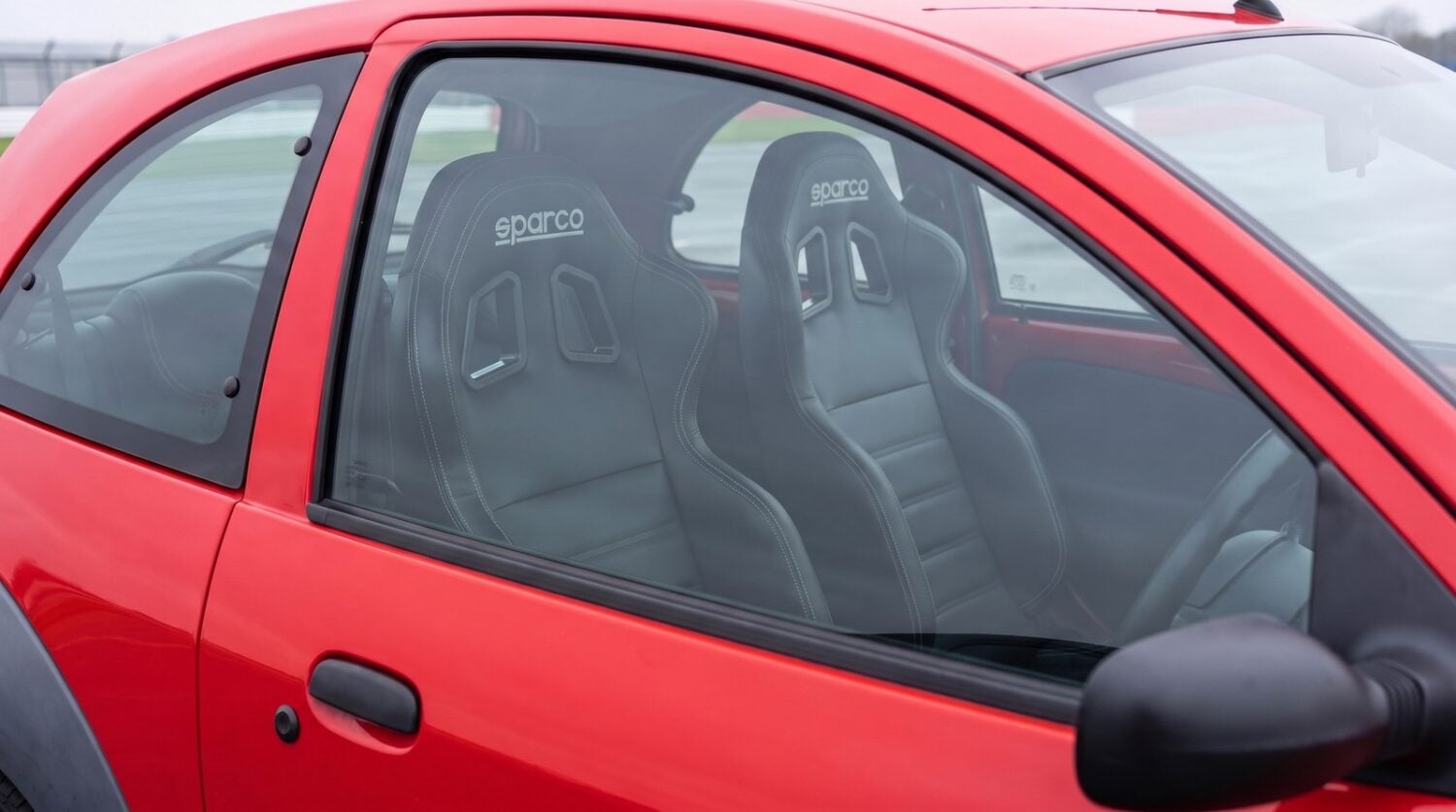Pair of black Sparco R100 Vinyl reclining sports seats fitted in a Radiant Red Ford Ka Mk1, seen through the closed driver door window