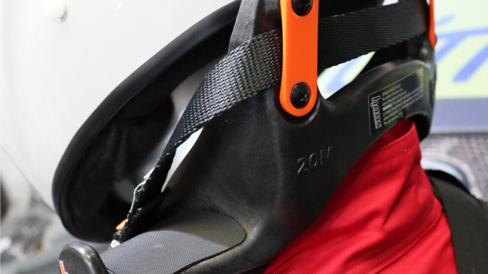 A HANS head and neck restraint worn with a white helmet and red race suit, its black tether running to the helmet