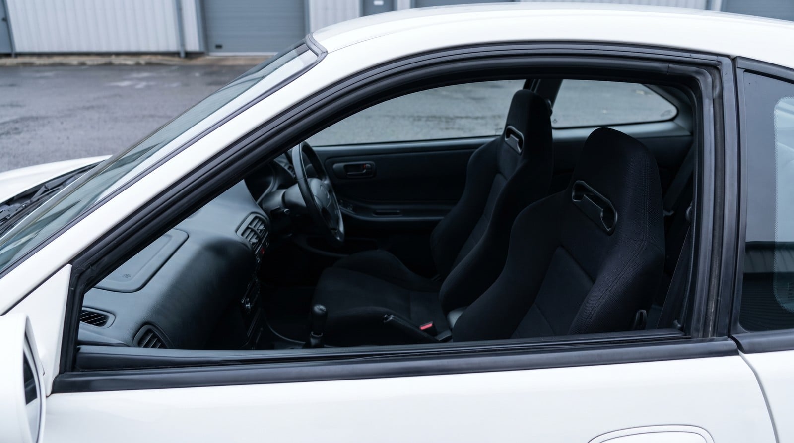Black bucket seats fitted in a white Honda Integra Type R DC2, seen over the open driver door window on an overcast day