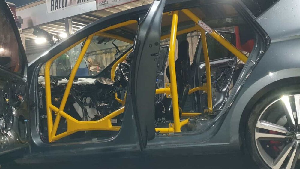 A yellow JP Cages competition roll cage with door bars fitted in a stripped grey Seat Leon MK3 five door, seen through the open front and rear doors