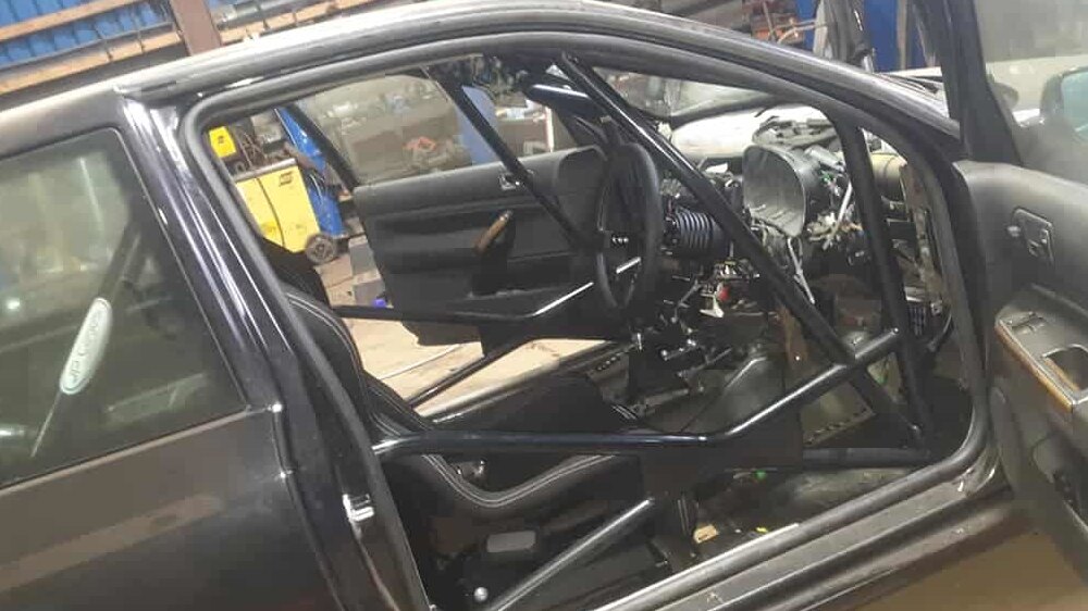 A black JP Cages bolt-in multipoint roll cage with door bars fitted in a stripped VW Golf MK4, seen through the open driver's door in a workshop