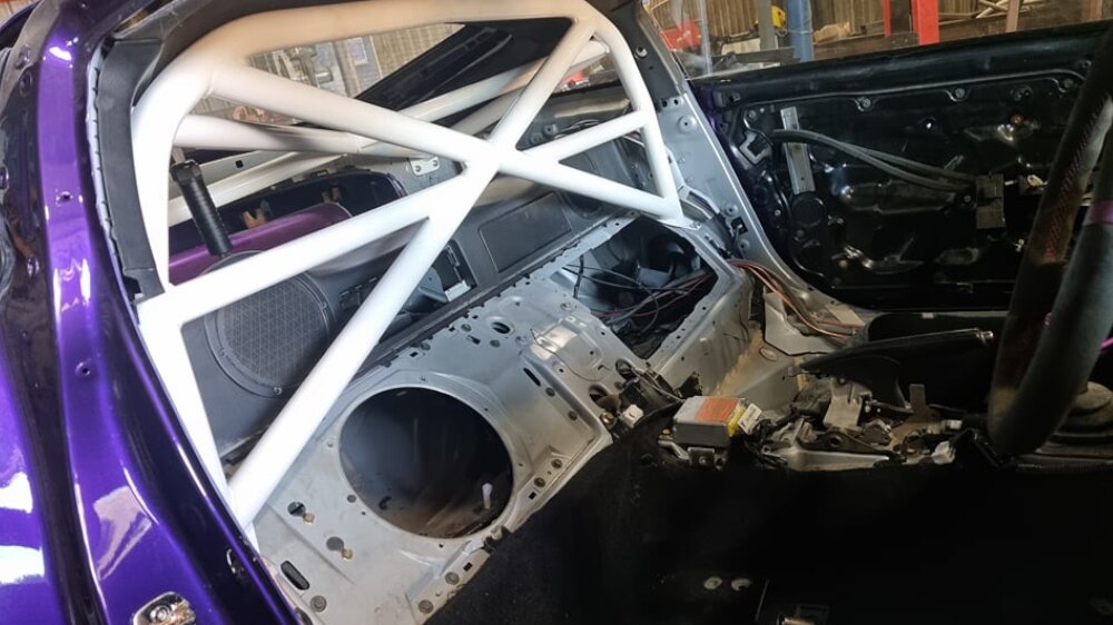 A white JP Cages show cage with X bracing and a harness bar fitted behind the seats of a purple Nissan 350Z with its interior stripped