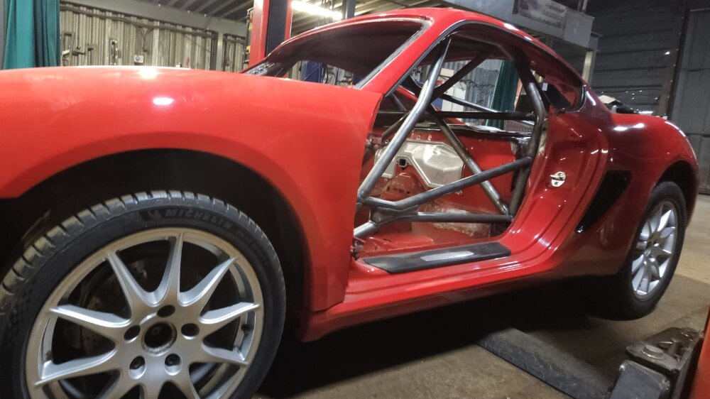 A red Porsche 987 shell in a workshop with a bare metal JP weld-in multipoint cage visible through the door opening