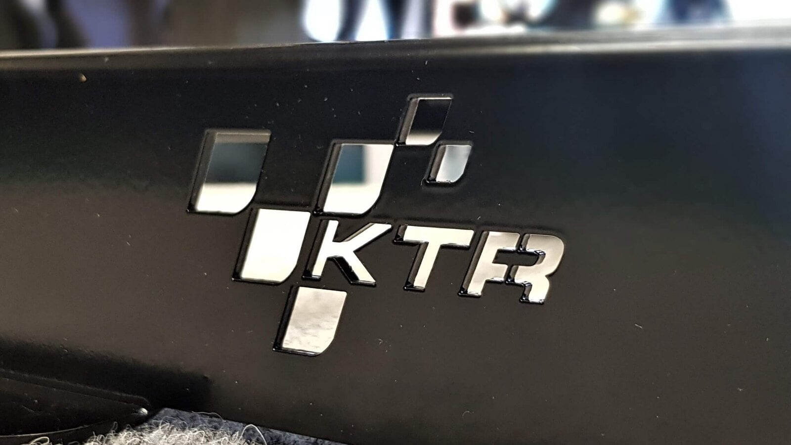 Close up of a black KTR Racing seat subframe with the cut out KTR logo, resting on grey carpet
