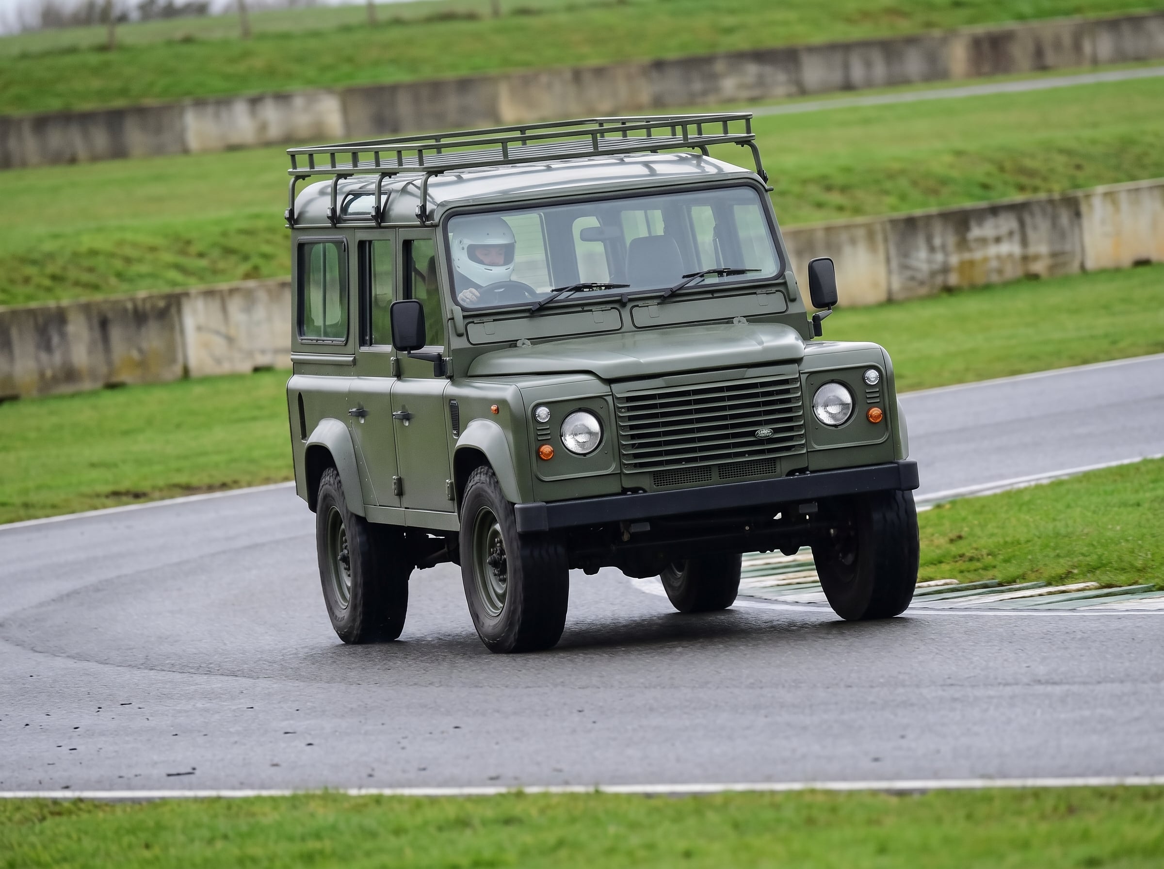 Classic Land Rover Defender 110 cornering on a British race circuit with helmeted driver visible through the windscreen