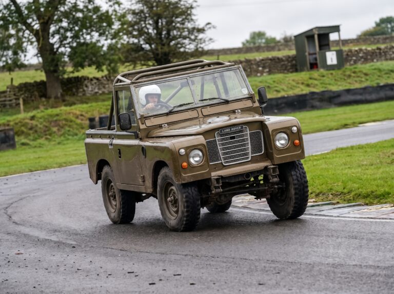 Bronze Green Land Rover Series 3 88-inch wheelbase cornering on a British race circuit with helmeted driver visible