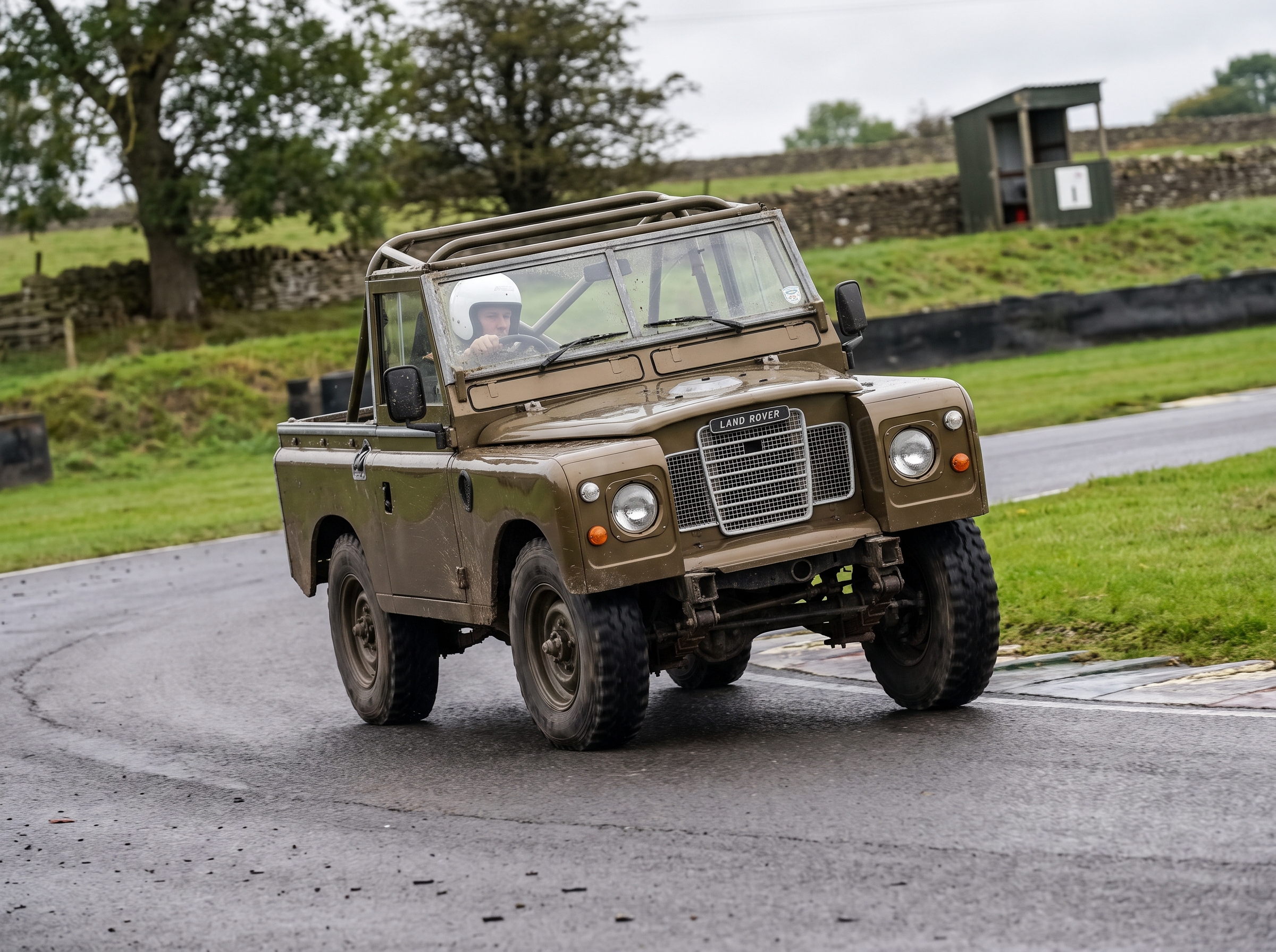 Bronze Green Land Rover Series 3 88-inch wheelbase cornering on a British race circuit with helmeted driver visible