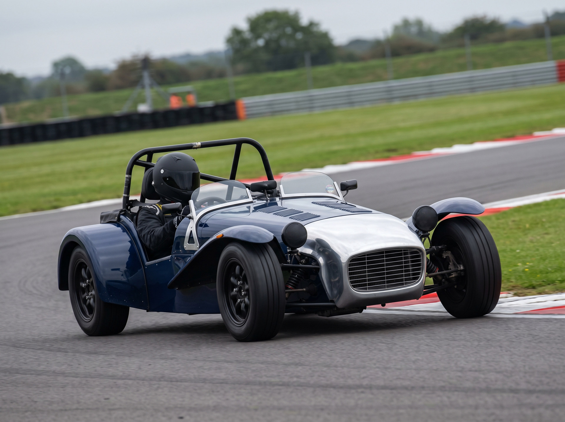 A dark blue Seven style kit car with a helmeted driver cornering on a British race circuit