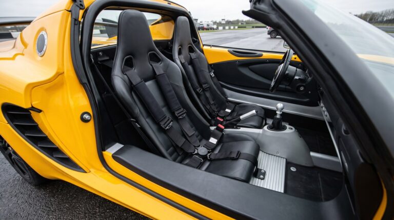 A black four point racing harness laid over the bucket seats of a yellow Lotus Elise with the roof removed, seen over the closed driver door