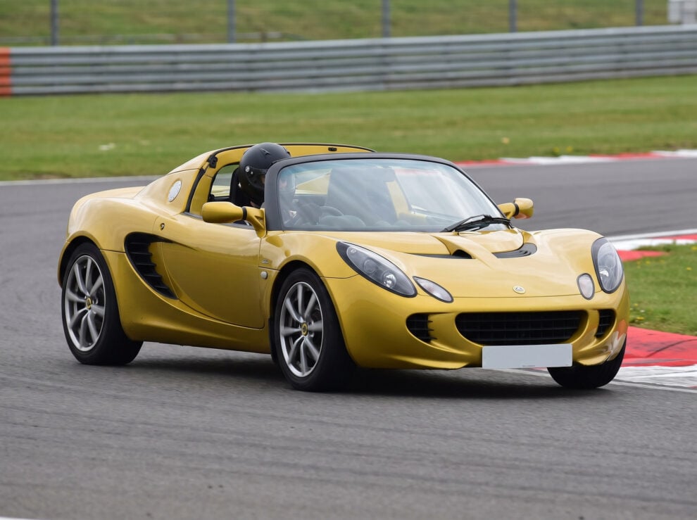 A yellow Lotus Elise S2 with a helmeted driver cornering on a British race circuit
