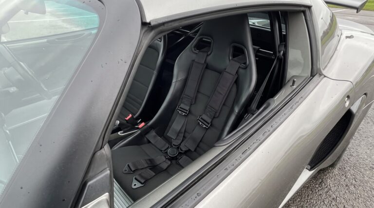 A black four point racing harness laid over the bucket seat of a grey Lotus Exige with a roll cage bar behind, seen over the closed driver door